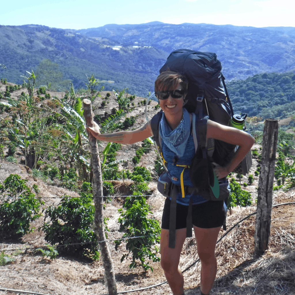 A young woman stopped to smile while hiking, leaning against a wooden fence post with a very large backpack on.