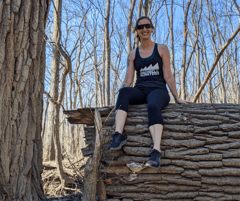 A smiling young woman sitting on a fallen tree while hiking. She's ready to share her beginner hiking tips.