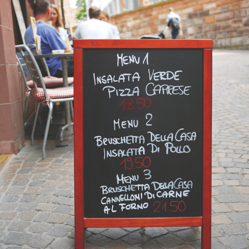 Outdoor blackboard near people sitting outdoors on cafe tables with chalk written dinner menu options.